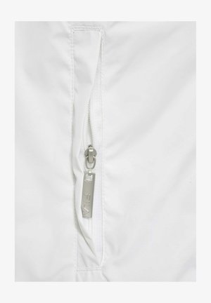 Balta jaka ar rāvējslēdzēja kabatu un sudraba rāvējslēdzēju, ar teksturētu audumu un smalkiem šuvumu detaļām.