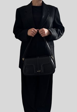 Persona con indosso un completo nero che tiene una borsa in pelle nera strutturata con doppi manici su uno sfondo grigio uniforme.