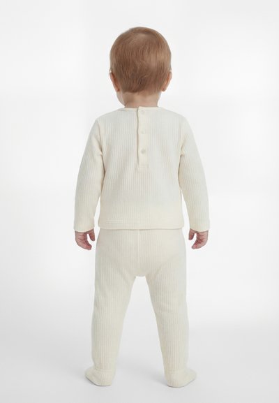 Heledate juustega väikelaps seisab seljaga, seljas kreemikat värvi soonikkoes pikkade varrukatega pluus, millel on nupud seljal, ja sobivad püksid.