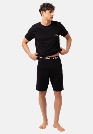 Jeune homme soulevant un t-shirt noir FILA, portant un short noir assorti FILA avec une ceinture à logo, debout pieds nus sur un fond uni.