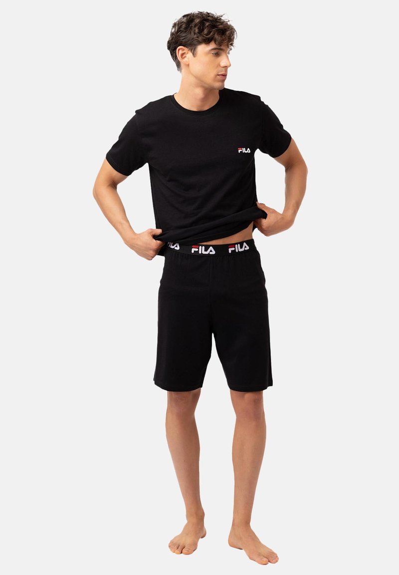 Jeune homme soulevant un t-shirt noir FILA, portant un short noir assorti FILA avec une ceinture à logo, debout pieds nus sur un fond uni.