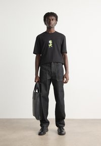 Camiseta negra de gran tamaño con gráfico de dinosaurio verde, combinada con pantalones oscuros de mezclilla de cintura alta y zapatos negros, sosteniendo una bolsa de tela negra.