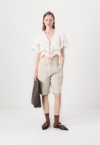 Hvit, kort topp med hullmønster og knytting, kombinert med beige skreddersydde shorts. Tilbehør inkluderer en brun veske og mørke slip-ons.