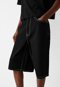 Schwarze Denim-Shorts mit weißer Steppung, entspannter Passform, schrägen Fronttaschen und knielanger Schnitt. Schlichtes Design.