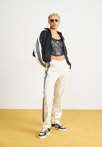 Svart och vit jacka i träningsstil med en kortärmad svart konstlädertröja, krämfärgade byxor med sidostråk och svarta sneakers med gula snören.