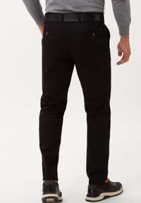 Pantaloni neri in cotone con una vestibilità slim, dotati di tasche posteriori con bottoni e un design semplice. Abbinati a scarpe da ginnastica nere.