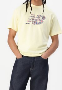 Osoba z warkoczami, ubrana w jasnożółtą koszulkę z kolorowym abstrakcyjnym wzorem oraz ciemnoniebieskie dżinsy, z rękami w kieszeniach.