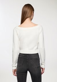 Maglione bianco lavorato a maglia con ampia scollatura off-the-shoulder, maniche lunghe e polsini a coste; abbinato a jeans attillati grigi scuri.