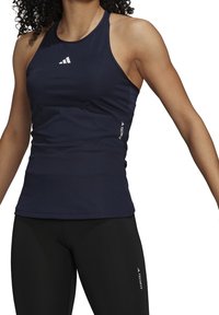 Žena v námořnickém bezrukávovém sportovním topu a černých legínách, oboje s malými bílými logy "Techfit" a Adidas.