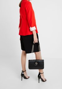 Červené sako s bílými manžetami, černé šortky a černé lakované podpatky. Drží černou kabelku s krokodýlím vzorem a řetízkovými popruhy.