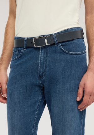 Homme portant un jean en denim bleu et une ceinture en cuir noire avec une boucle argentée, debout les mains détendues le long du corps.