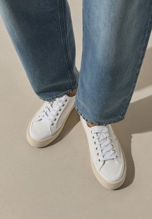 Pies con zapatillas blancas con cordones y vaqueros azules, de pie sobre un suelo de color claro con sombras.