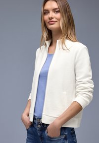 Junge Frau trägt eine weiße, strukturierte Jacke mit Reißverschluss über einem hellblauen Oberteil und blauen Jeans, steht mit den Händen in den Taschen vor einem grauen Hintergrund.