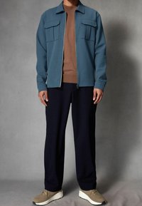 Lichtblauwe jas met rits en twee borstzakken, daaronder een bruine lange mouwen shirt, donkere broek en beige sneakers. Soepele stoftextuur.