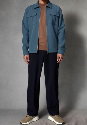 Hellblauer Reißverschlussjacke mit zwei Brusttaschen, darunter ein braunes langärmeliges Hemd, dunkle Hose und beigefarbene Turnschuhe. Glatte Stoffstruktur.