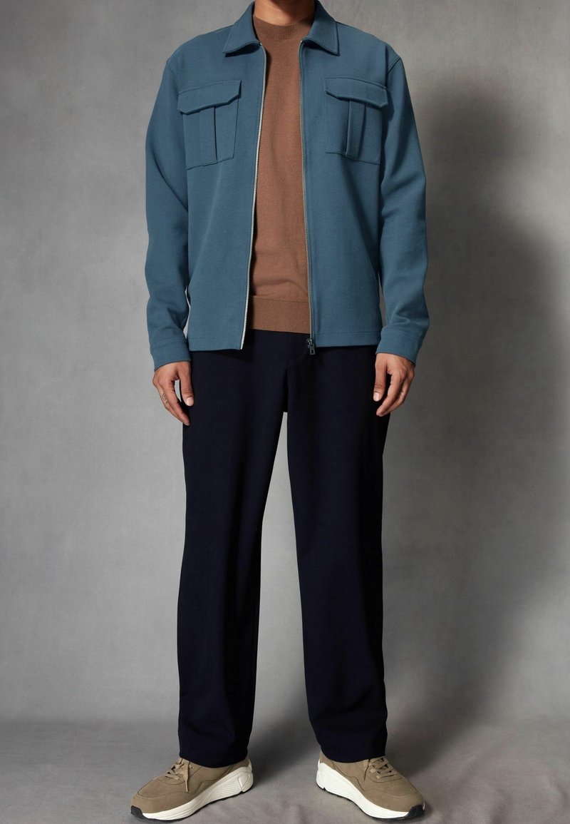 Lichtblauwe jas met rits en twee borstzakken, daaronder een bruine lange mouwen shirt, donkere broek en beige sneakers. Soepele stoftextuur.