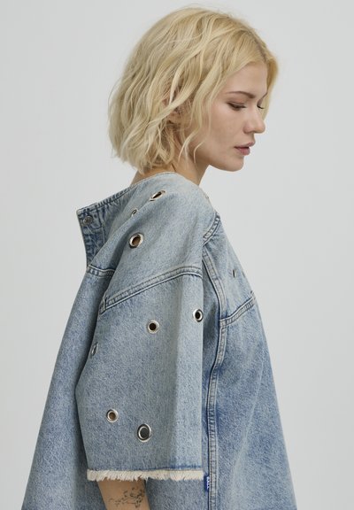 Femme blonde de profil portant une grande veste en jean claire avec des œillets en métal et des bords de manches effilochés sur un fond uni.