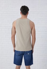 Beige ærmeløs tanktop lavet af blødt stof, parret med blå denim shorts med frynser. Baggrunden er en hvid murstensvæg.