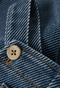 Lähedane vaade sinisest denimkangast, kus on oranž õmblus ja ümmargune, nelja auguga beež nupp, mis on õmmeldud beežide niitidega.