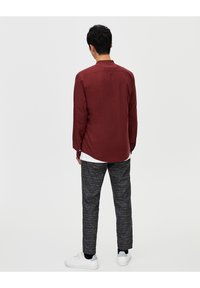 Chemise rouge à manches longues avec des détails de boutons au col, associée à un pantalon foncé à motifs et des baskets blanches, vue de dos.