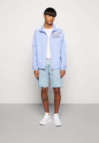 Lyseblå hættejakke med hvide striber, stort Puma-logo. Båret med hvid t-shirt og slidte denimshorts; parret med hvide sneakers.