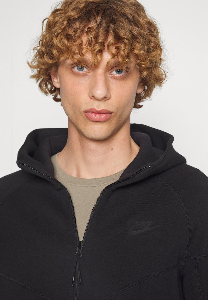 Svart zip-up hoodie med en texturerad finish, med en Nike-logotyp på bröstet och en grå crew neck-tröja under.