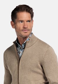 Beige zip-up trui met een textured breisel en een hoge kraag. Daaronder voegt een blauw bloemenhemd een contrast toe.