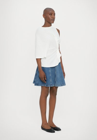 Biely krátky rukáv s uzlovým detailom v kombinácii s rozšírenou riflovou sukňou. Čierne baleríny dopĺňajú outfit. Hladká textúra látky.