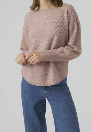 Mujer con suéter de punto texturizado color malva con patrón en zigzag y jeans azules, de pie con las manos entrelazadas al frente.