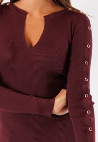 Top a maniche lunghe rigato color bordeaux con scollatura a V profonda. Presenta dettagli in pizzo sulle maniche. Realizzato in tessuto morbido e elasticizzato.