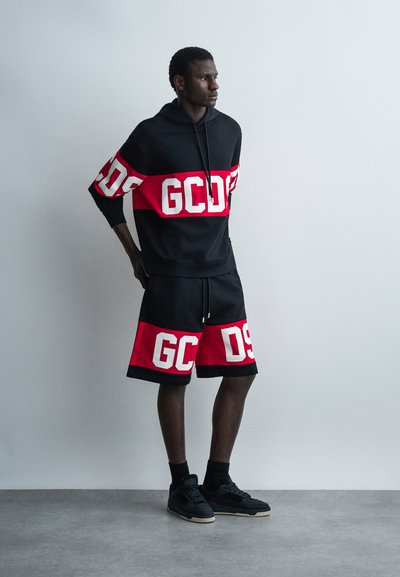 Giovane uomo in piedi con felpa con cappuccio nera e rossa e pantaloncini coordinati con lettere bianche a blocchi, indossa scarpe da ginnastica nere davanti a uno sfondo semplice.