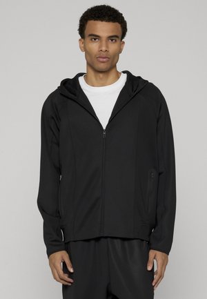 Fiatal férfi fekete cipzáras kapucnis dzsekit visel fehér póló fölött, világos szürke egyszínű háttér előtt áll.