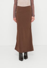 Jupe marron moulante, longue jusqu'au sol, en tissu lisse et mat, associée à des bottes noires à bout pointu et un haut transparent à manches longues.