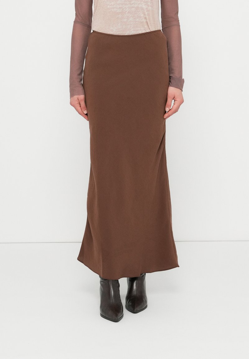 Jupe marron moulante, longue jusqu'au sol, en tissu lisse et mat, associée à des bottes noires à bout pointu et un haut transparent à manches longues.