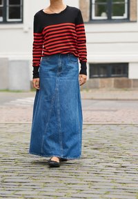 Camisa de manga larga a rayas rojas y negras combinada con una falda maxi de mezclilla azul. Zapatos planos negros, de pie sobre pavimento de adoquines.