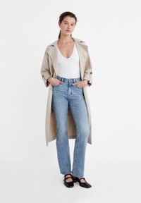 Beżowy płaszcz trench z białą bluzką, jasnoniebieskie jeansy z wysokim stanem i czarne płaskie buty. Prosty design bez widocznych wzorów czy detali metalowych.