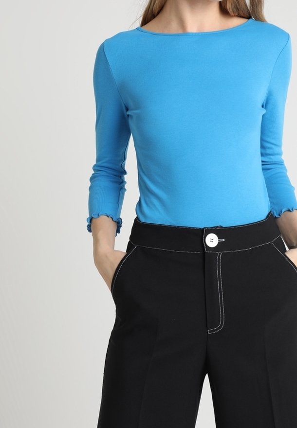 Långärmad, figurnära topp i livfull blå med volangdetaljer vid ärmsluten. Matchad med högsittande svarta byxor med vit söm och knapp.