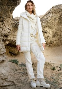 Witte pufferjack met capuchon, gestructureerde trui, witte broek en beige laarzen met veters. Outfit tegen een rotsachtige, zandige achtergrond.