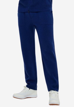 Marineblaue Strickhose mit Fronttaschen, getragen mit weißen Low-Top-Sneakers vor weißem Hintergrund.