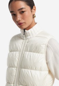 Gepolstertes Zip-Up-Weste in Off-White, ausgestattet mit einem hohen gerippten Kragen und glatter Textur. Enganliegend mit gesteppten Abschnitten und einem eleganten Design.