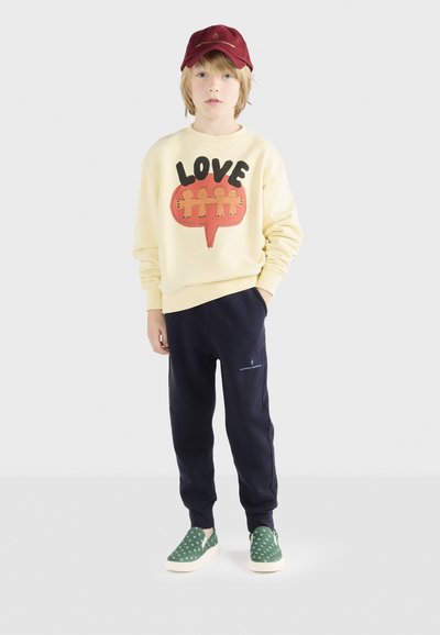 Gelbes Sweatshirt mit "LOVE"-Grafik, marineblaue Jogginghose und grüne Slipper mit weißen Punkten. Eine rote Baseballkappe rundet das Outfit ab.