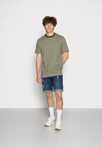 T-shirt listrado em verde-azeitado com decote redondo, combinado com calções de ganga e ténis brancos com detalhes laranja; modelo posicionado contra um fundo neutro.