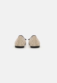 Beige Ballerinas mit einem gesteppten Muster, schwarzem Rand und einer Schleifendetail. Flache Gummisohlen. Die Rückansicht zeigt eine abgerundete Ferse und eine glatte Textur.