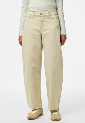 Person iført beige bukser med vide ben, lysegrå bluse med lange ærmer og beige sneakers, stående på hvid baggrund.
