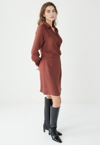 Brązowa, długa sukienka z długim rękawem, zapinana na krótko z kołnierzykiem, dopasowana w talii i o gładkiej fakturze, połączona z czarnymi, skórzanymi kozakami do kolan.