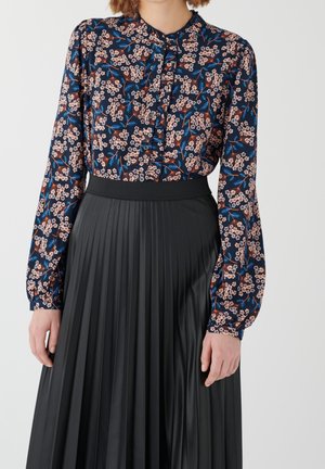 Femme portant un chemisier bleu marine à manches longues avec un motif floral rose, rentré dans une jupe plissée noire taille haute, sur un fond uni.