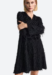 Fekete ruha bolyhos francia hatású textúrával, V-nyakkivágással, hosszú ujjal és laza szabással. Térd alá érő szegély, egyenetlen élekkel és enyhe fényességgel.