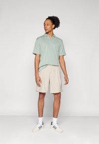 Polo verde claro de manga curta com um padrão de losangos, combinado com calções beges. O modelo usa ténis brancos com detalhes em preto.