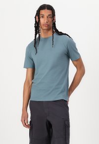 T-shirt de algodão azul-petróleo com gola redonda e mangas curtas, apresentando um corte descontraído e uma textura suave, usado com calções cargo escuros.
