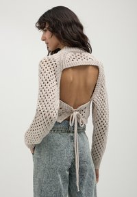 Donna che indossa un maglione lavorato a maglia beige con schiena scoperta, legato in vita, abbinato a jeans leggeri a vita alta blu chiaro, in piedi contro uno sfondo neutro.